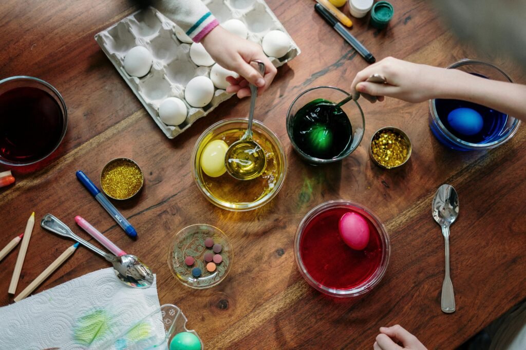 Hands dip eggs into bowls of food coloring on a craft-filled table