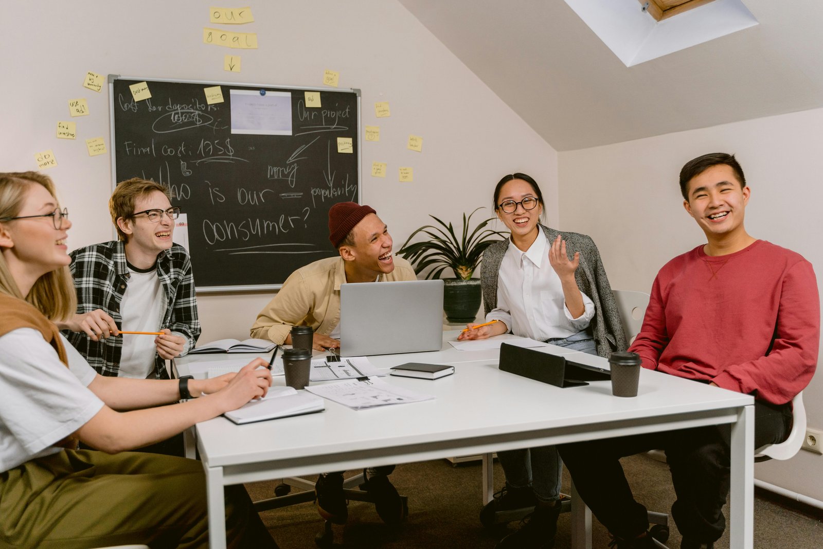 A diverse group of young professionals engaged in a lively discussion around a table, brainstorming ideas with notes on a blackboard in the background, related to EEO classifications.